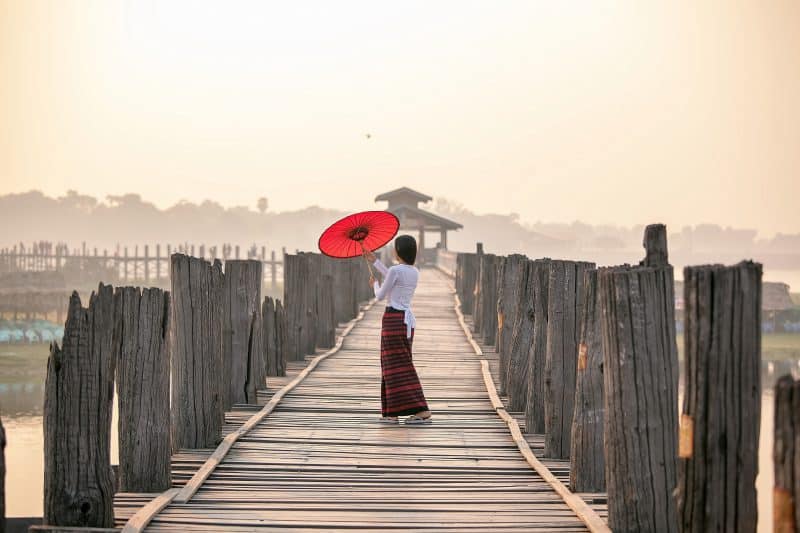 668965135 - ผู้หญิงกำลังถือร่มสีแดง. เดินบนสะพานอูเบ็ง
